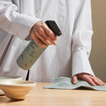 Guests on Earth non-toxic cleaning products: hand spraying refillable cleaner and wiping a table beside ceramic bowls