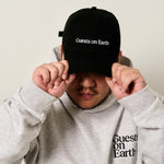 Guests on Earth black ambassador cap with white logo, worn by model adjusting the brim.