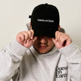 Guests on Earth black ambassador cap with white logo, worn by model adjusting the brim.