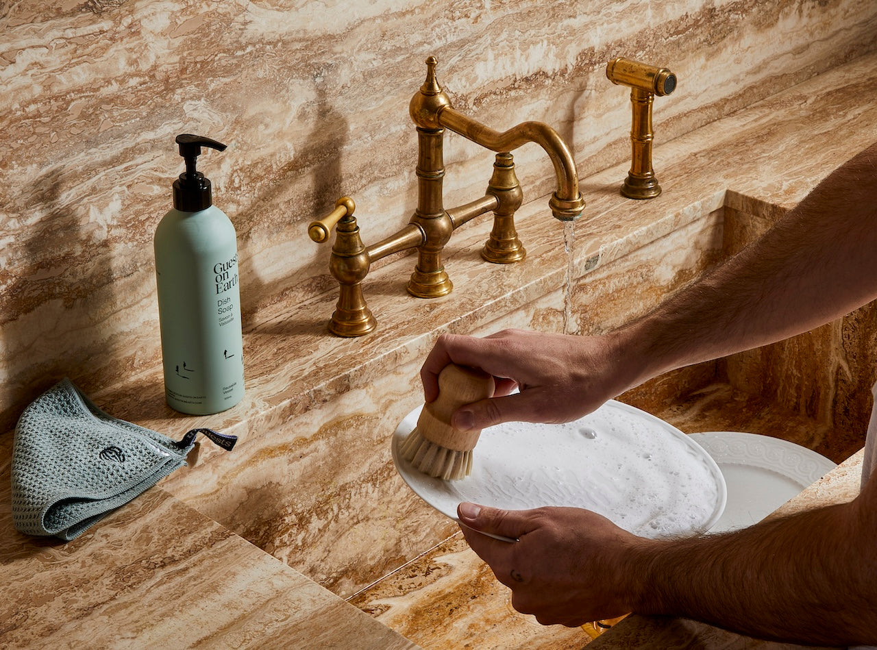 Guests on Earth dish soap beside a soapy sink, as a plate is scrubbed under warm brass fixtures.