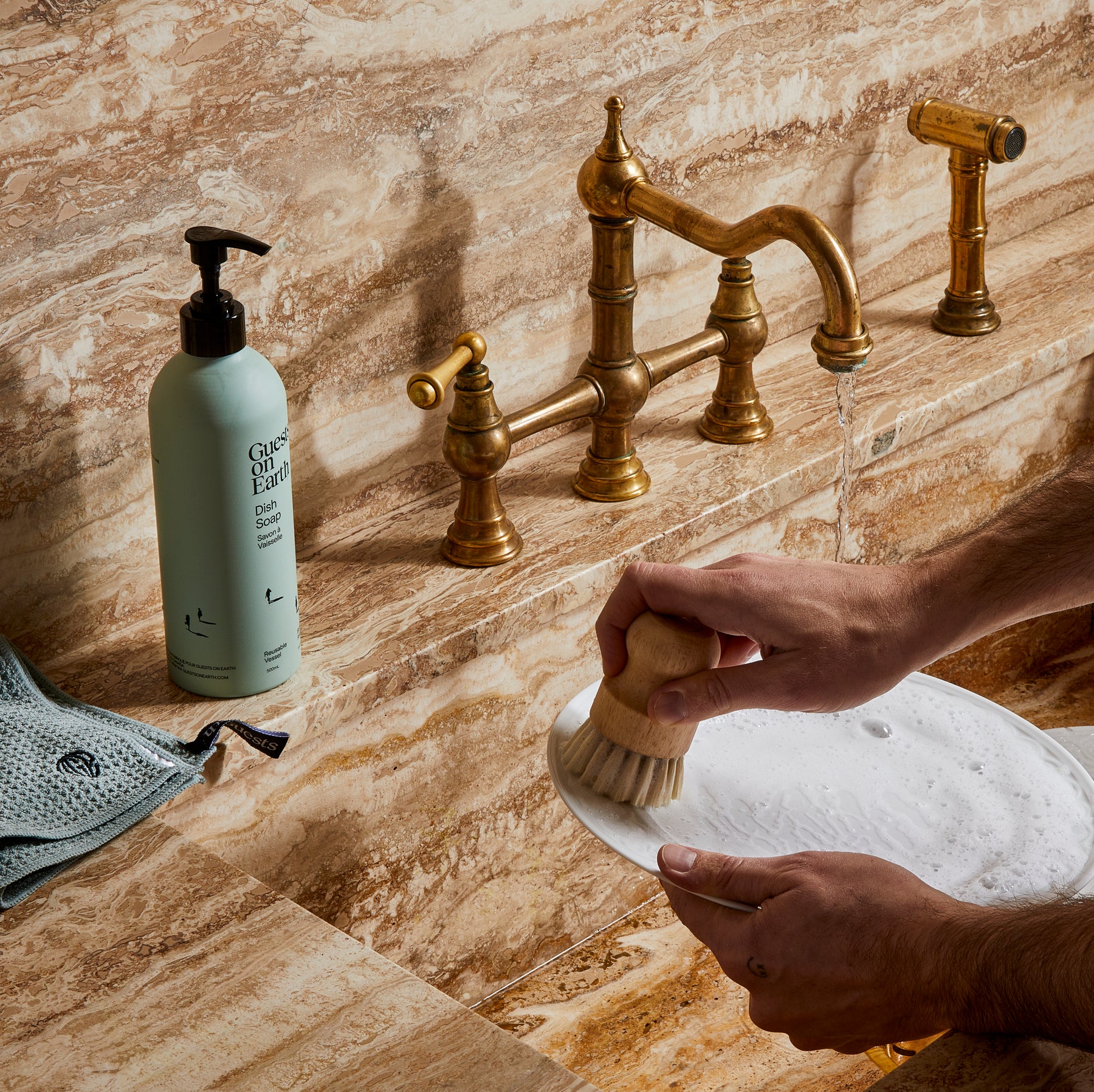 Guests on Earth Dish Soap by the marble sink as hands scrub a plate with a wooden brush under running water