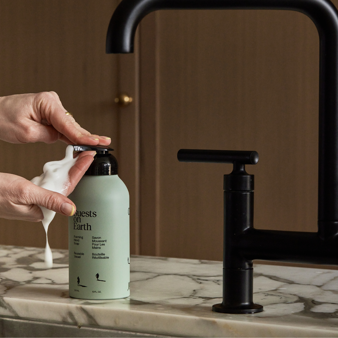 Guests on Earth Foaming Hand Soup dispensed into hands at a marble sink, sleek refillable bottle beside matte-black tap