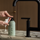 Guests on Earth Foaming Hand Soup dispensed into hands at a marble sink, sleek refillable bottle beside matte-black tap